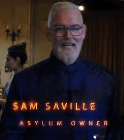 A man with a beard and glasses wearing a dark, ornate shirt, smiling as text overlays state his name 'Sam Saville' and his title 'Asylum Owner'.