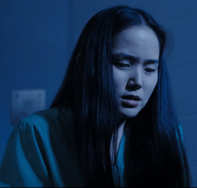 A woman with long black hair wearing a teal garment, looking down with a concerned expression in a dimly lit space.